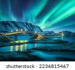 Bridge and northern lights over snowy mountains. Lofoten islands, Norway. Aurora borealis and reflection in water. Winter landscape with starry sky, polar lights, rocks, road, sea, city illumination