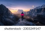 Small photo of Woman on the rock and mountain peaks at night in autumn in Dolomites, Italy. Girl on the stone, high rocks, sky with moon, light trails on road at twilight in fall. Colorful landscape with cliffs