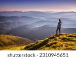 Girl on mountain peak with green grass looking in beautiful mountain valley in fog at sunset in autumn. Landscape with sporty young woman, foggy hills, forest, sky in fall. Travel and tourism. Hiking