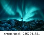 Aurora borealis and young woman on mountain peak at night. Northern lights, stones and silhouette of alone girl on mountain trail. Landscape with polar lights. Starry sky with bright aurora. Travel