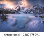 Frozen sea coast and rocks in snow at colorful sunset. Winter in Lofoten islands, Norway. Snowy mountain peaks, lake with frosty shore, ice, trees, colorful sky with clouds golden light. Landscape