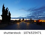 Scenery of Charles Bridge before sunrise with street lamp lights glowing in blue twilight & silhouettes of majestic statues & towers of historical buildings under dawning sky in Old Town Prague, Czech