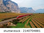 Small photo of Aerial view of a fast train dashing through the beautiful landscape of vineyard fields on a sunny autumn day and a rugged mountain wall dominating the background, in Mezzocorona, Trentino, Italy