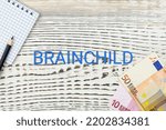 Small photo of BRAINCHILD - word (text) and euro money on a white wooden table, notebook, notepad. Business concept: buying, selling, commerce (copy space).