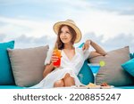 Young romantic woman in straw hat holding mai tai sits by table with alcoholic blue lagoon cocktail in martini glass at outdoors bar, restaurant at luxury resort, hotel. Relax, rest, holiday, vacation