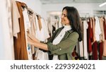 Small photo of Happy woman stands in a fashion store, carefully choosing clothing items to buy. She browses through racks of stylish clothes, examining each one closely before making a decision.