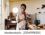 Small photo of Brazilian woman holding a coffee cup and looking away thoughtfully in her home kitchen. home. Mature black woman standing by a breakfast table filled with freshly baked cheese bread rolls.