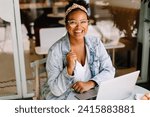 Young woman sits in a café, working on her laptop. She is a freelancer and content writer, enjoying the freedom of remote work. With a smile, she embodies the happiness of a successful entrepreneur.