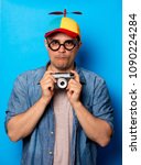 Small photo of Young nerd man with noob hat holding a vintage photo camera on blue background