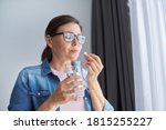 Mature middle-aged woman in casual clothes at home holding pill and glass of fresh water. Headaches, depression, medicine supplements vitamins, sedatives, menopause