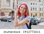 Fashionable smiling happy beautiful girl teenager 16, 17 years old with wireless headphones with bright dyed colored hairstyle on street of summer sunny city. Lifestyle, youth, fashion, beauty