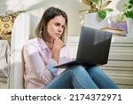 Serious sad young woman looking at laptop screen, sitting on floor at home