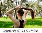Young brunette woman wearing sportive clothes on city park, outdoors tying her hair in a ponytail while doing some workout.