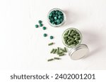 Two glass jars filled with capsules and tablets of organic spirulina on a white textured background. Top view. Natural cleansing of the body