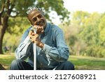 Small photo of Aging man having joint pain in his knee in a lush green park in noon. Elderly retired man holding a walking stick. Showcasing health care Osteoarthritis concept. Old man showing various expressions.