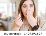 Beautiful young woman applying red lipstick cover mouth with hand shocked with shame for mistake, expression of fear, scared in silence, secret concept
