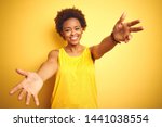 Beauitul african american woman wearing summer t-shirt over isolated yellow background looking at the camera smiling with open arms for hug. Cheerful expression embracing happiness.