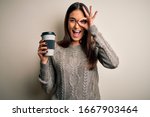Young beautiful brunette woman drinking glass of coffee over isolated white background with happy face smiling doing ok sign with hand on eye looking through fingers