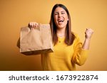 Young beautiful woman holding take away paper bag from delivery over yellow background screaming proud and celebrating victory and success very excited, cheering emotion