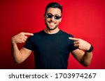 Small photo of Young handsome man wearing funny thug life sunglasses over isolated red background looking confident with smile on face, pointing oneself with fingers proud and happy.