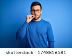 Young handsome man with beard wearing casual sweater and glasses over blue background mouth and lips shut as zip with fingers. Secret and silent, taboo talking