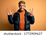 Young handsome skier man with beard wearing snow sportswear and ski goggles showing and pointing up with fingers number seven while smiling confident and happy.