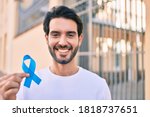 Small photo of Young hispanic man smiling happy holding blue ribbon at the city.