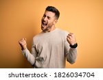 Young handsome man wearing casual sweater standing over isolated yellow background very happy and excited doing winner gesture with arms raised, smiling and screaming for success. Celebration concept.