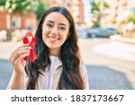 Young hispanic woman smiling happy holding red hiv ribbon walking at the city.