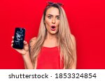 Young beautiful blonde woman holding broken smartphone showing cracked screen scared and amazed with open mouth for surprise, disbelief face