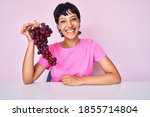 Beautiful brunettte woman holding branch of fresh grapes looking positive and happy standing and smiling with a confident smile showing teeth 