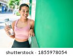 Young beautiful hispanic sporty woman wearing fitness outfit smiling happy and natural leaning on a green chroma wall at the town drinking fresh water from the bottle