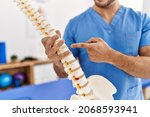 Young hispanic man wearing physio therapist uniform pointing to anatomical model of vertebral column at clinic