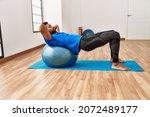 Handsome hispanic man doing exercise and stretching on yoga mat, practicing flexibility with pilates ball at the gym