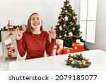Small photo of Caucasian young blonde woman sitting on the table by christmas tree gesturing finger crossed smiling with hope and eyes closed. luck and superstitious concept.
