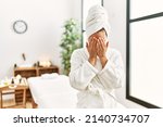 Young brunette woman wearing towel and bathrobe standing at beauty center with sad expression covering face with hands while crying. depression concept. 