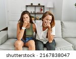 Small photo of Mother and daughter together sitting on the sofa at home smelling something stinky and disgusting, intolerable smell, holding breath with fingers on nose. bad smell
