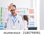 Middle age hispanic man working at pharmacy drugstore surprised with hand on head for mistake, remember error. forgot, bad memory concept. 