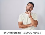 Small photo of Hispanic man with beard standing over isolated background with hand on chin thinking about question, pensive expression. smiling with thoughtful face. doubt concept.