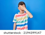 Small photo of Young caucasian kid standing over blue background covering eyes with hand, looking serious and sad. sightless, hiding and rejection concept