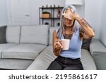 Small photo of Middle age grey-haired woman drinking coffee sitting on the sofa at home covering eyes with hand, looking serious and sad. sightless, hiding and rejection concept