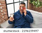 Hispanic young man wearing bathrobe and eye bags patches drinking wine speaking on the phone celebrating crazy and amazed for success with open eyes screaming excited. 