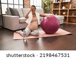 Young pregnant woman sitting on yoga mat with pilates ball smiling happy doing ok sign with hand on eye looking through fingers 
