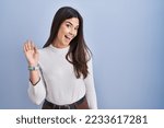 Small photo of Young brunette woman standing over blue background waiving saying hello happy and smiling, friendly welcome gesture