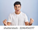 Small photo of Hispanic man standing over blue background angry and mad raising fists frustrated and furious while shouting with anger. rage and aggressive concept.