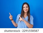 Small photo of Young brunette woman standing over blue background smiling and looking at the camera pointing with two hands and fingers to the side.