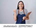 Young brunette woman wearing professional waitress apron and clipboard screaming proud, celebrating victory and success very excited with raised arms 