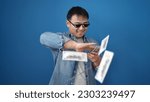Small photo of Young chinese man wearing thug life glasses throwing dollars over isolated blue background