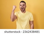 Small photo of Young hispanic man standing over yellow background angry and mad raising fist frustrated and furious while shouting with anger. rage and aggressive concept.