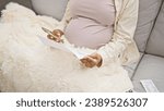 Small photo of Tucked in a blanket at home, a young, pregnant woman, warmly seated on her sofa, indulges in motherhood duties while reading important baby documents using her trusty smartphone technology.